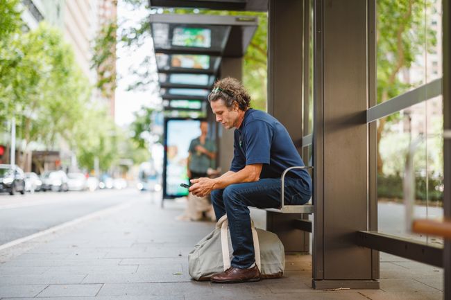 Man sat at a bus stop in the city looking down at his phone with a duffel bag between his legs
