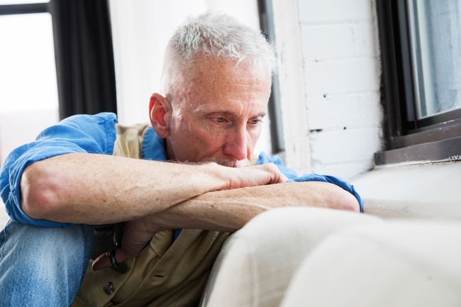 Man sat on a couch, one kneee up, covering the lower part of his face, looking into the wall